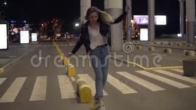 年轻的金发女人穿着牛仔裤和黑色皮夹克，晚上在黄色的街道保险杠上跳跃，玩得很开心。 街道